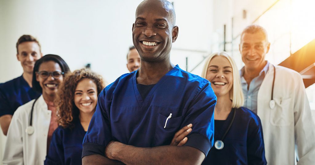 Young African male doctor smiling while standing in a hospital corridor with a diverse group of staff in the background; blog: 6 Solutions to Healthcare Staffing Challenges