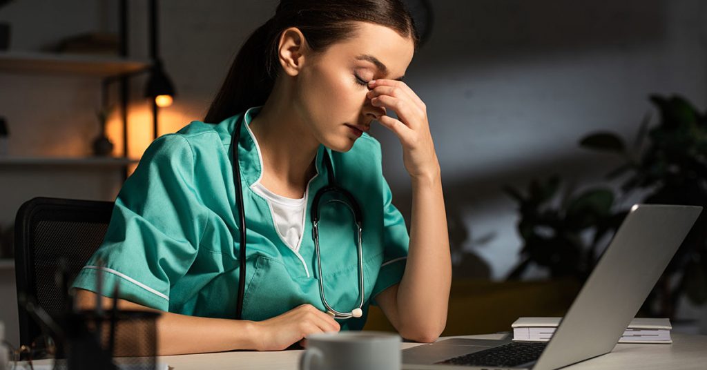 attractive and tired nurse in uniform sitting at table during night shift; How Technology Can Prevent Nurse Burnout